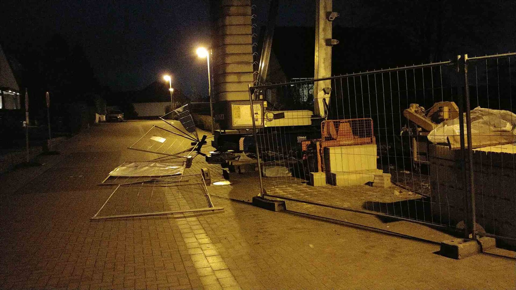 Nachtansicht einer Baustelle am Straßenrand mit Bauzäunen, Metallplatten und Kran.