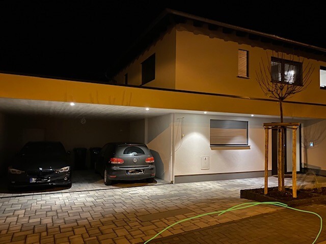 Nächtliche Wohnhausgarage mit zwei geparkten Autos, gepflastertem Vorplatz und Baum.