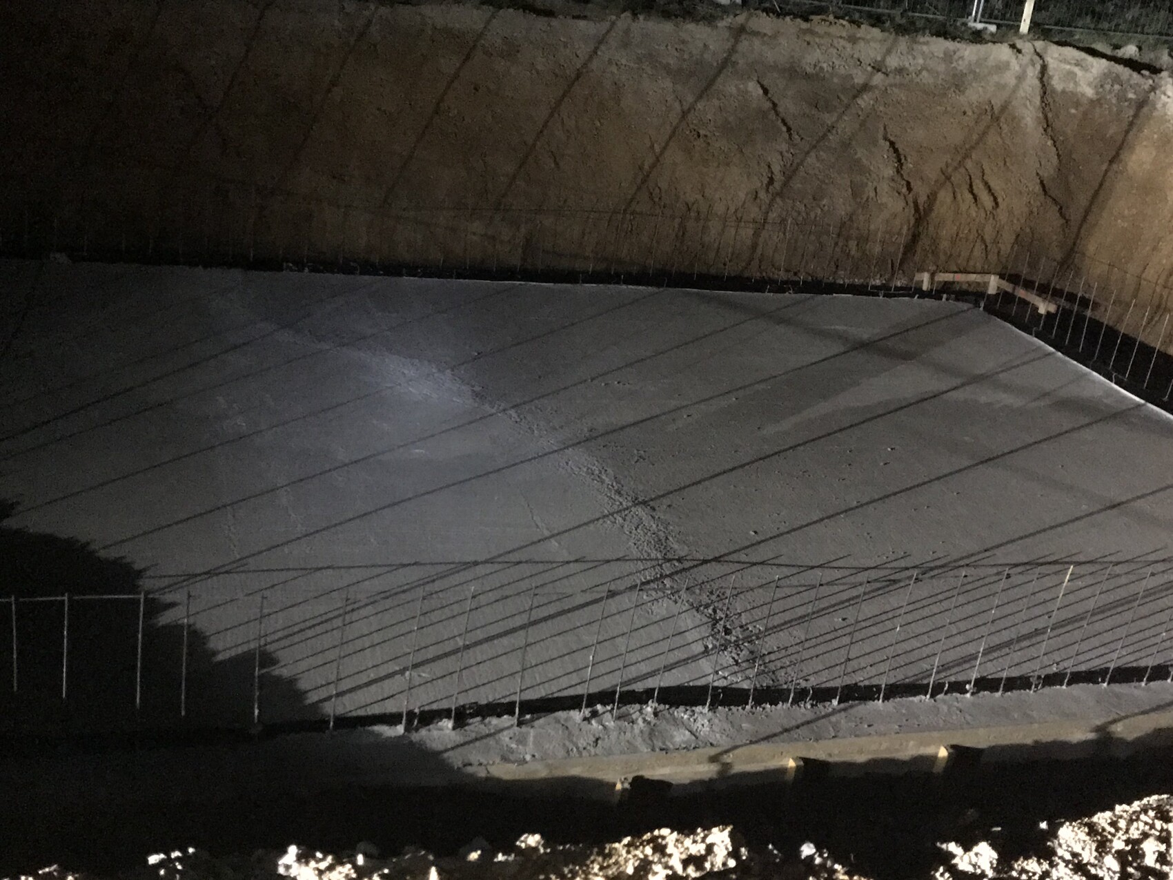 Nachtaufnahme einer Betonbodenplatte im Rohbau mit Bewehrung und Baugrube.