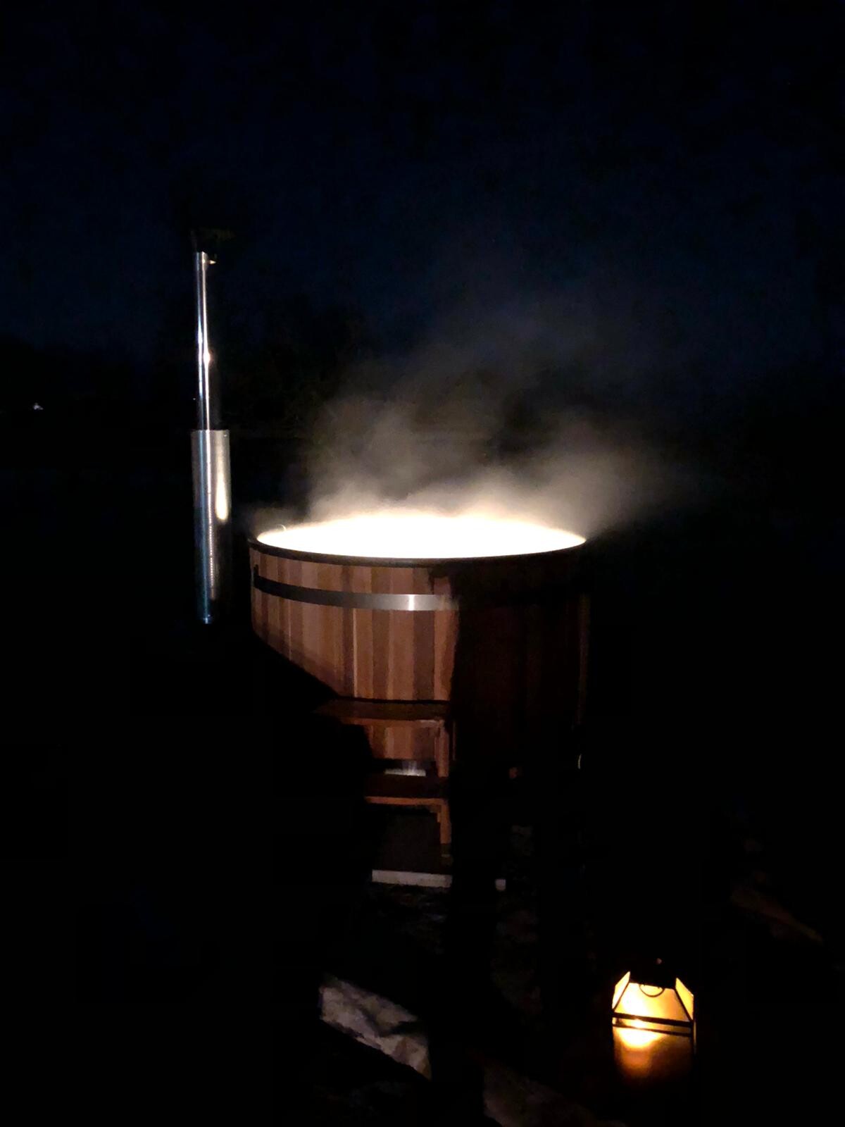 Nachtaufnahme: dampfende Holzbadewanne mit Kaminrohr und Laterne.