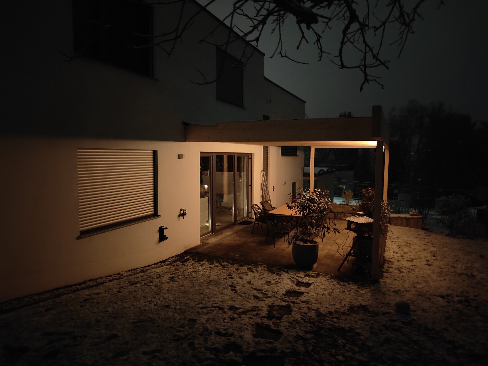 Nachts beleuchtete Terrasse eines modernen Hauses mit Esstisch und Stühlen; Schnee auf dem Boden.