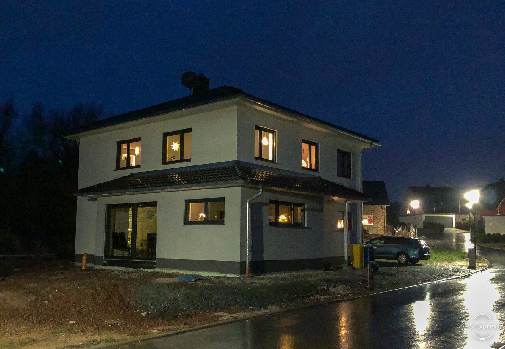 Beleuchtetes zweistöckiges Haus bei Nacht; warmes Licht aus den Fenstern, nasse Straße.