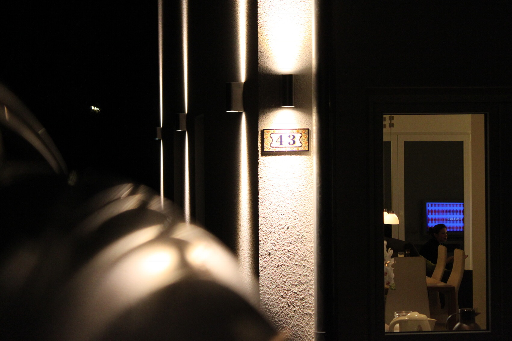 Nachtaufnahme der Gebäudefassade 43; Blick in beleuchteten Innenraum mit Person am Tisch.