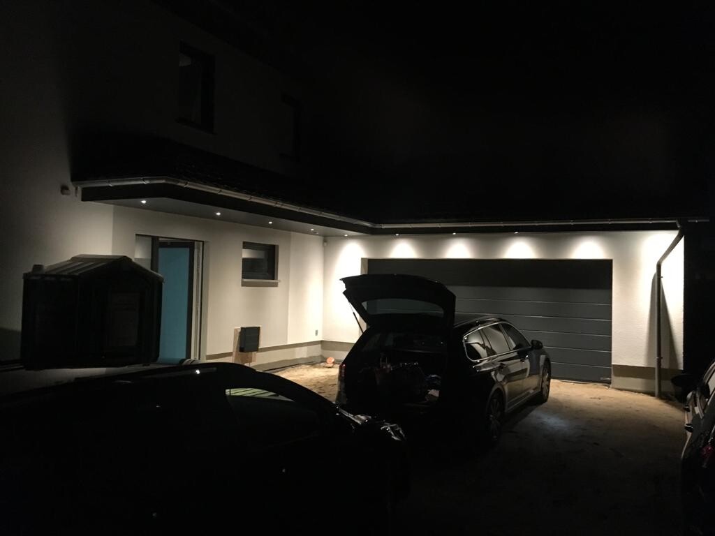 Nachts vor der Garage: Auto mit offenem Kofferraum, daneben weitere Fahrzeuge, Beleuchtung.