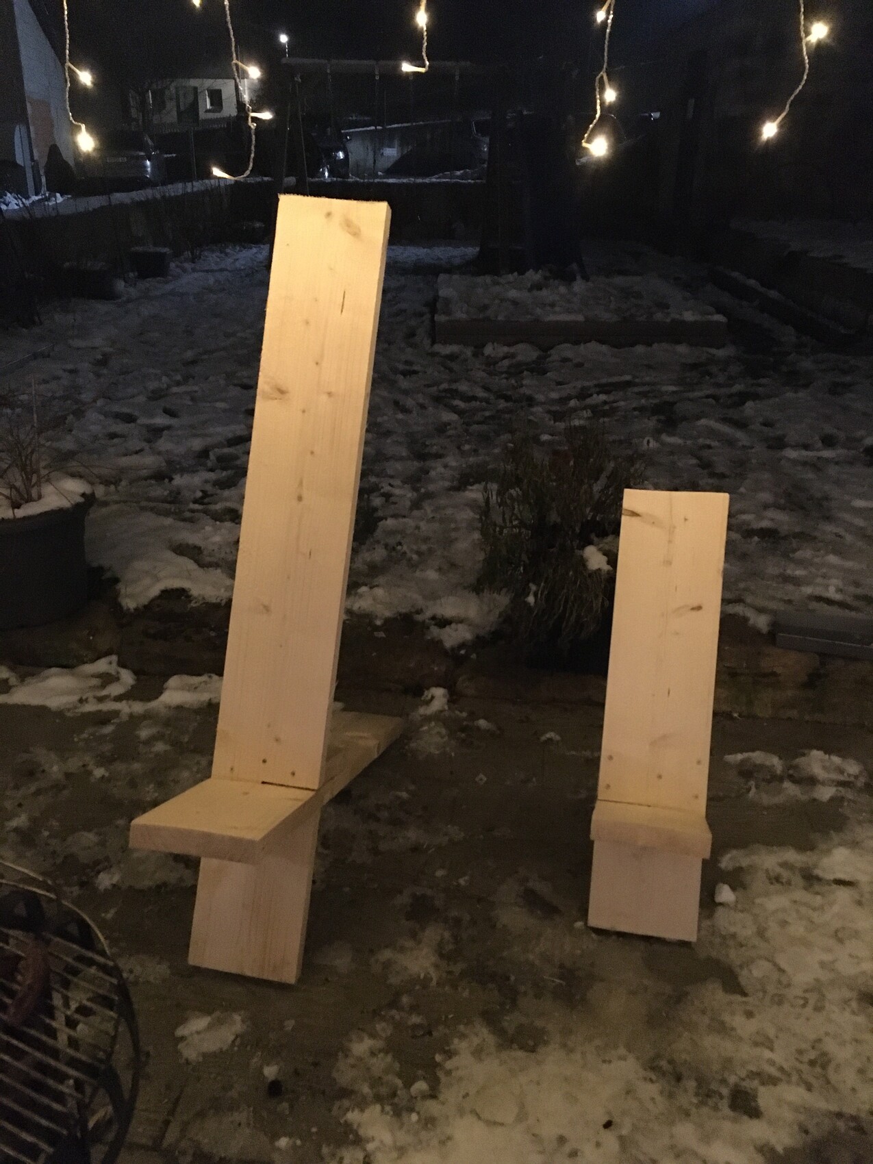 Zwei unbehandelte Holzplanken dienen als improvisierte Stühle im winterlichen Garten, Lichterketten.