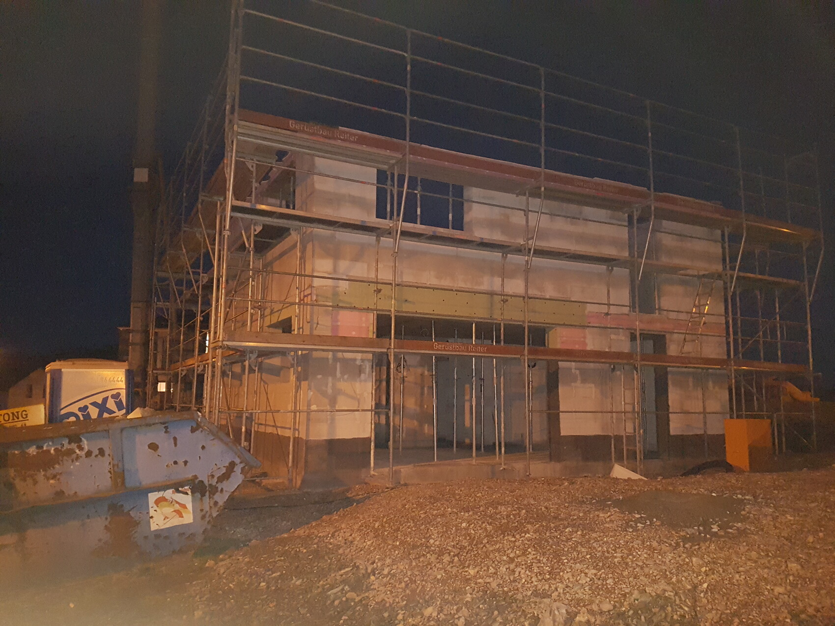 Baustelle bei Nacht: mehrstöckiges Gerüst um ein Gebäude, Kiesboden vorne, Müllcontainer links.