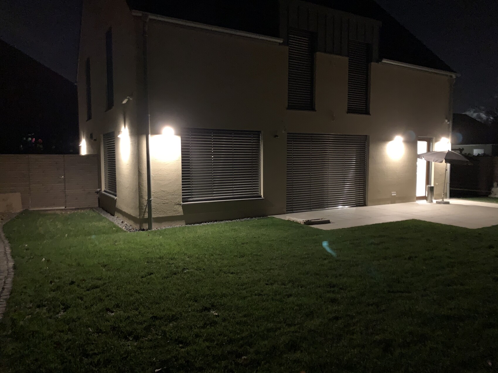 Nachtansicht eines modernen Hauses mit beleuchteten Außenwänden, großen Rollläden und Terrasse.