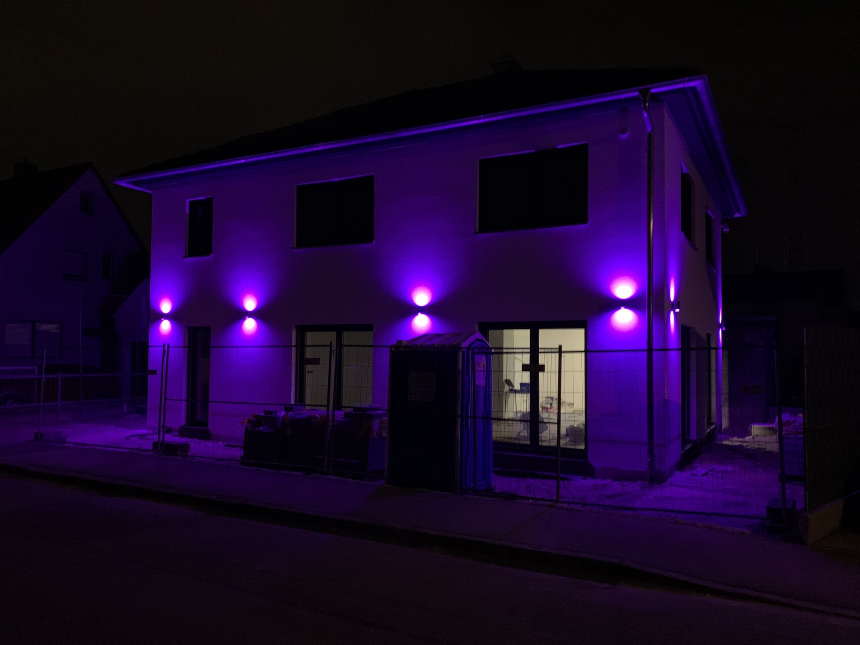 Haus bei Nacht mit violetten Außenleuchten an der Fassade und Bauzaun