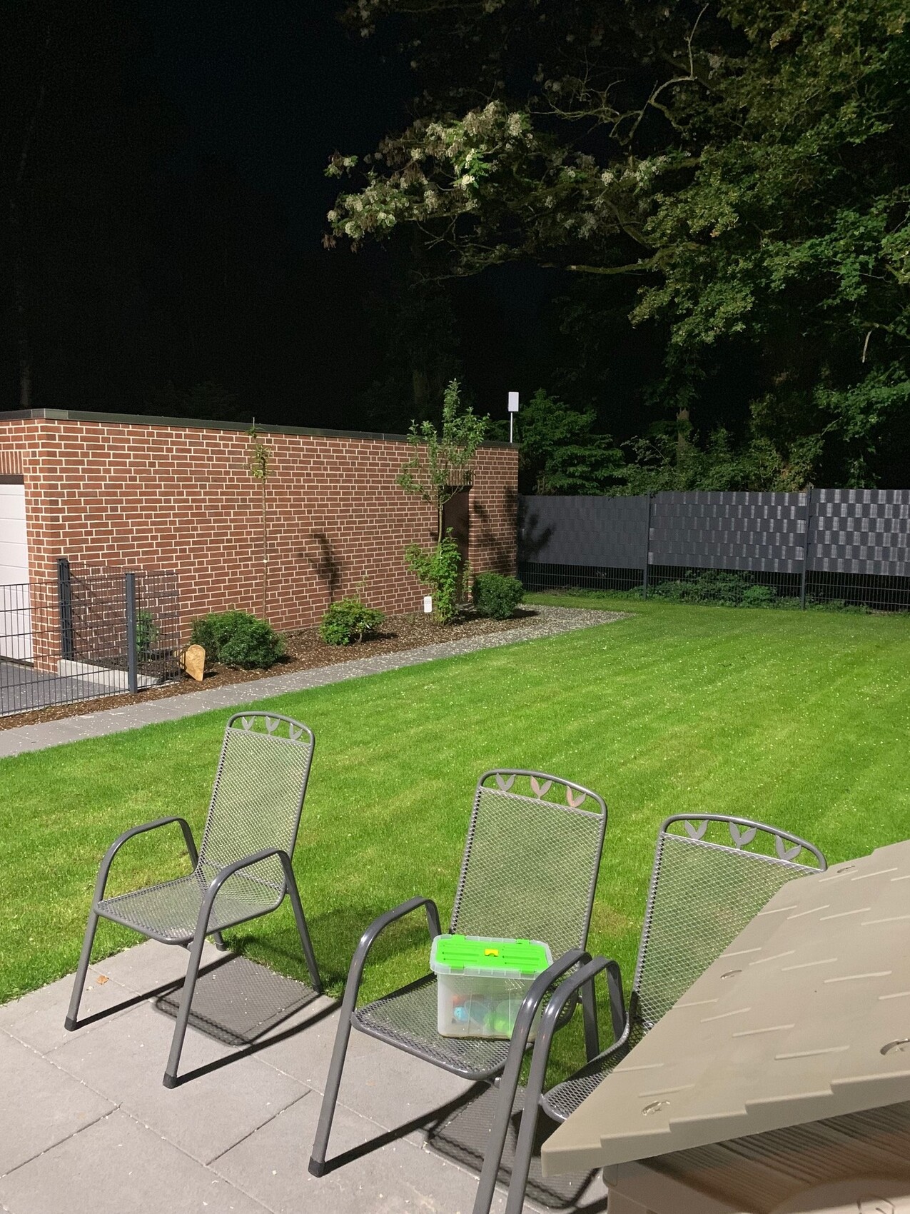 Nacht: drei Metallstühle auf Terrasse; grüne Werkzeugbox auf mittlerem Stuhl; Rasen dahinter.