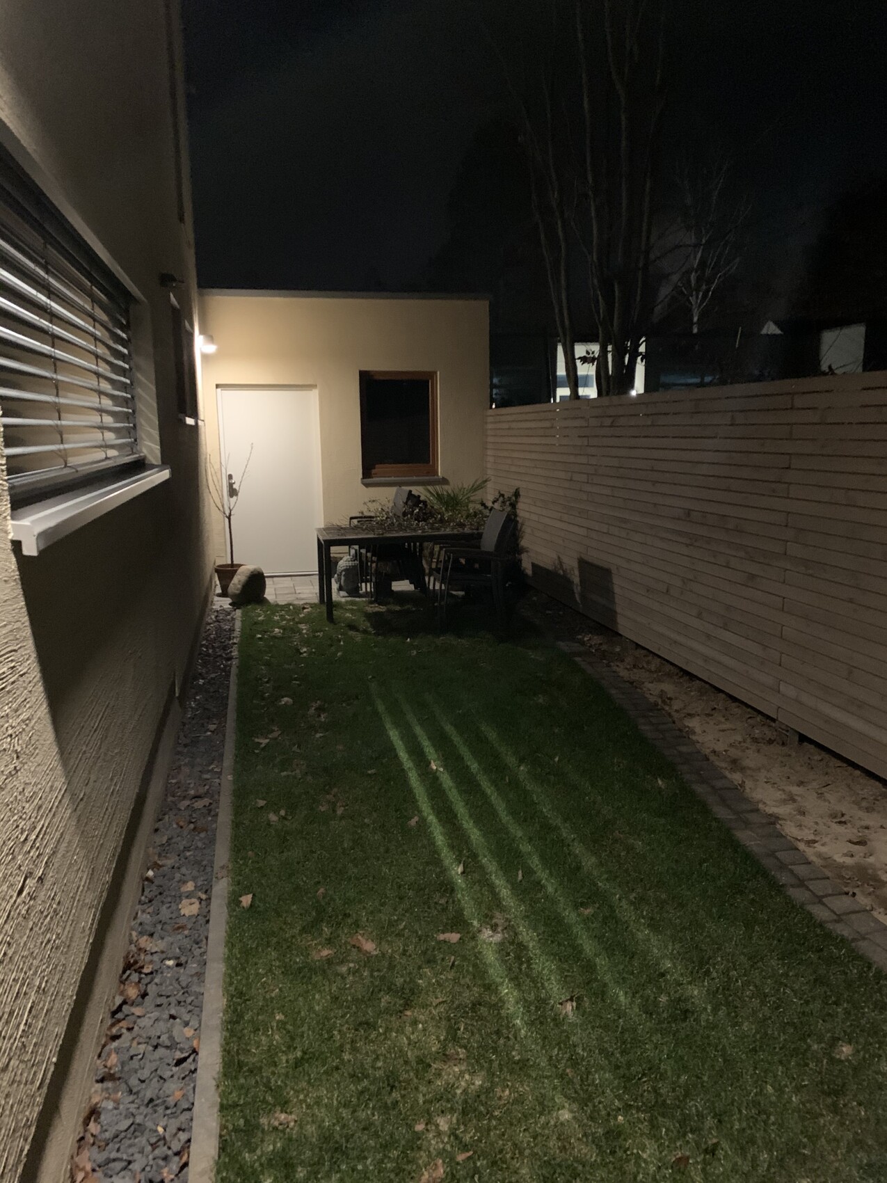 Nächtlicher Garten mit Tischgruppe, Tür, Fenster und Holzzaun an einer Seitenwand.
