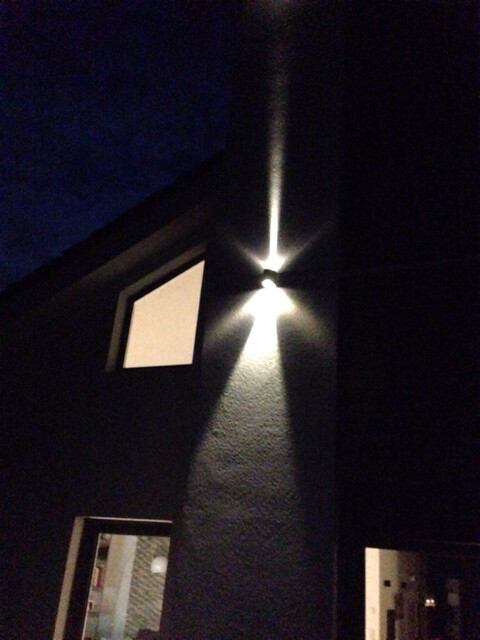 Nächtliche Außenansicht eines dunklen Gebäudes; Wandlampe wirft Lichtstrahl an die Fassade hinunter.