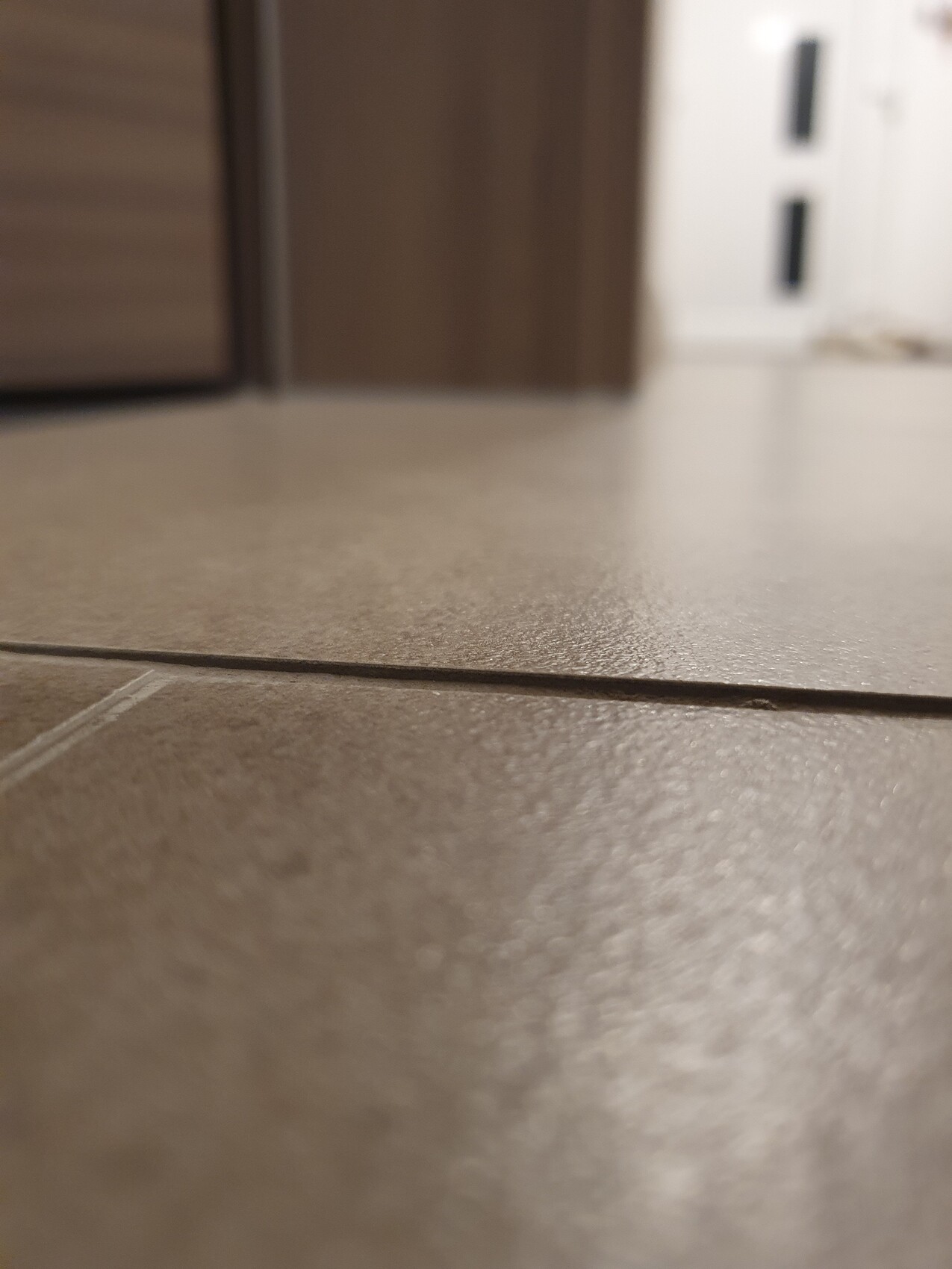 Nahaufnahme einer Bodenfliese mit Textur, Fuge im Vordergrund, unscharfer Hintergrund mit Möbeln.