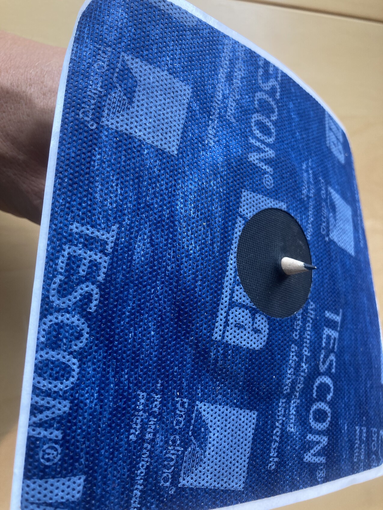 Nahaufnahme eines blauen, perforierten Stoffs mit schwarzer, runder Patch und Metallstift.