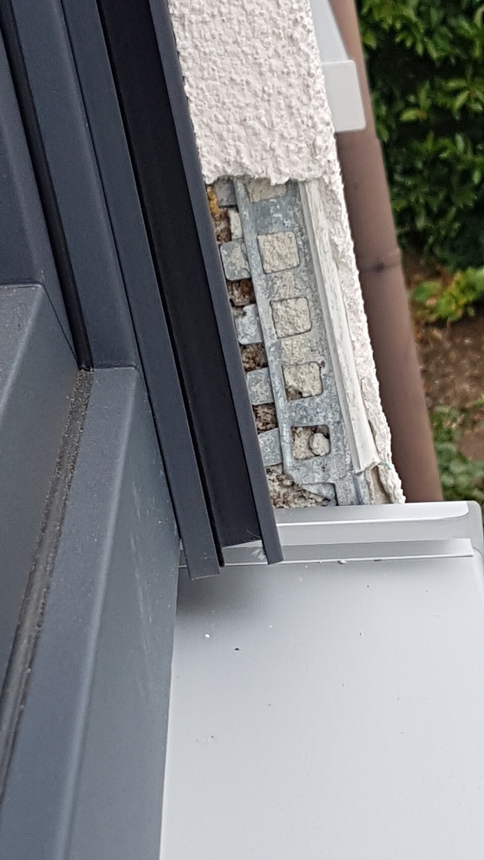 Außenwand mit dunklem Fensterrahmen, sichtbare Metallhalterung und Putzriss, grüne Hecke dahinter