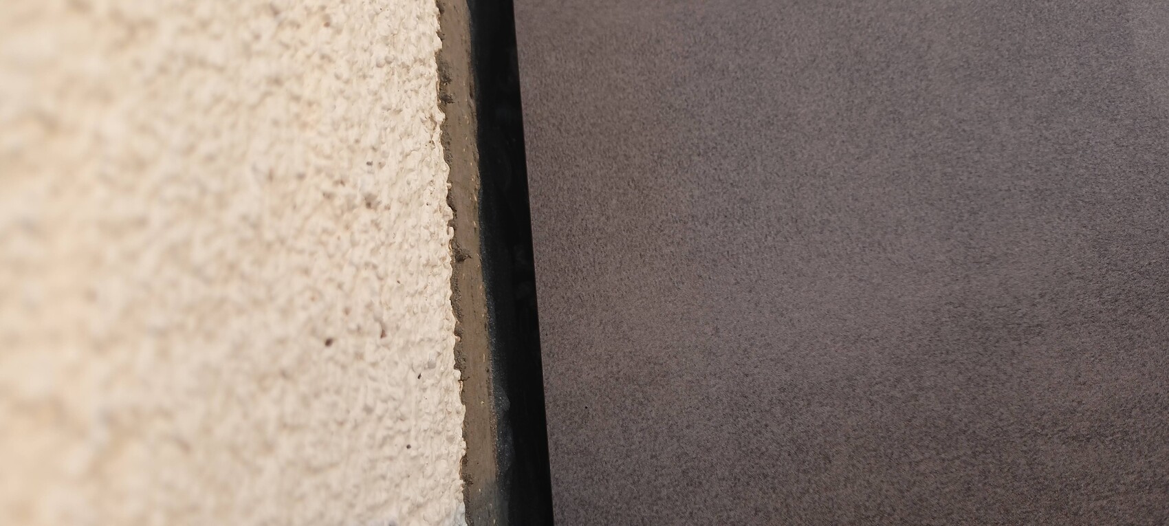 Nahaufnahme einer rauen hellbeigen Wand links, schmaler Spalt zu glatter dunkler Fläche rechts.
