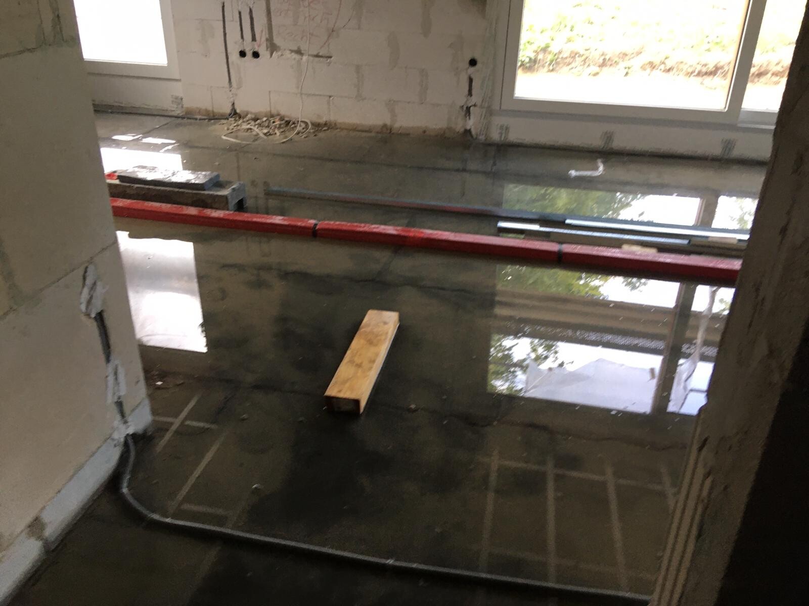 Baustellenraum mit stehendem Wasser auf dem Boden, roter Rohrleitung, Holzblock und Fenstern.
