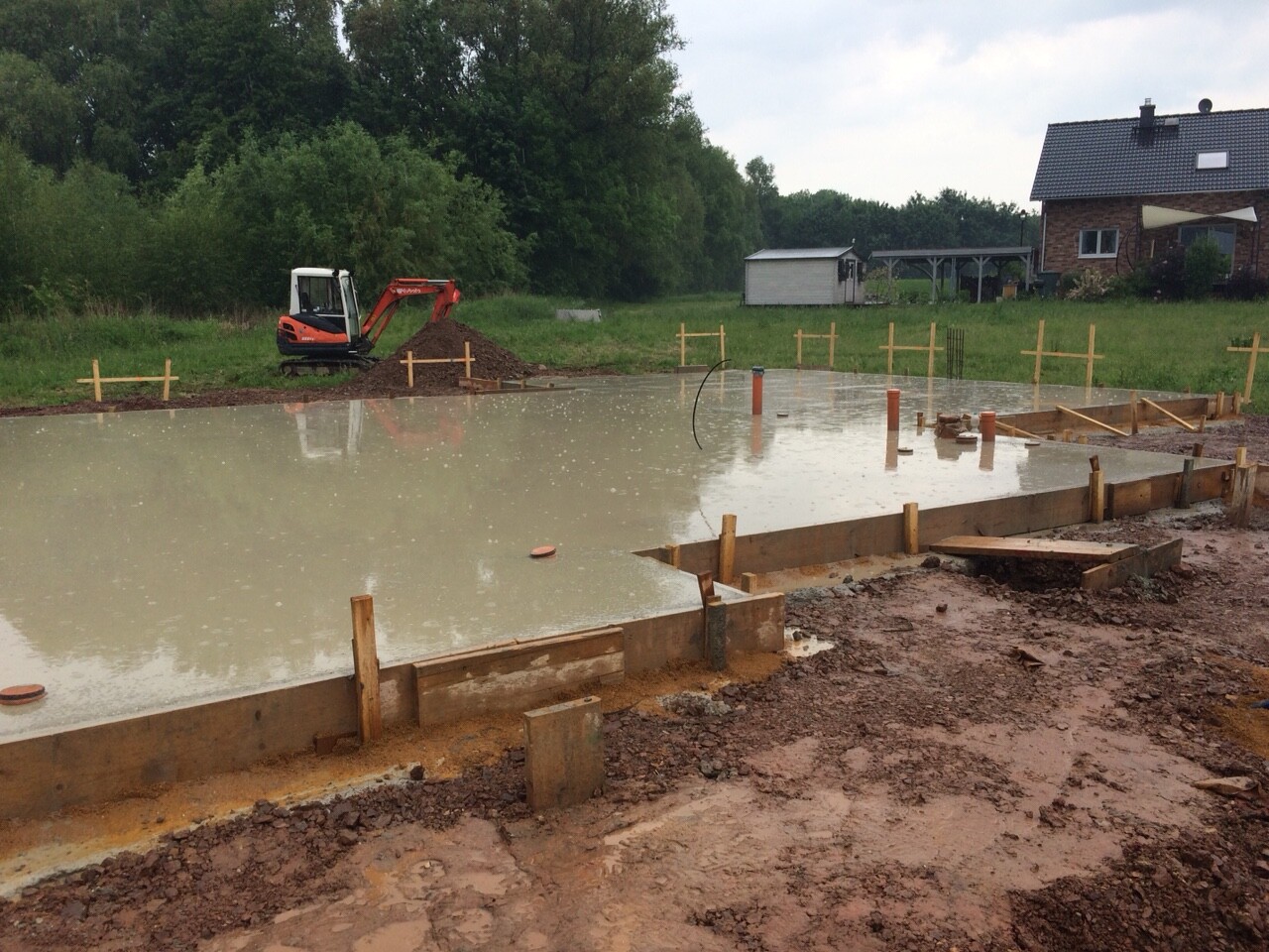 Frisch gegossenes Betonfundament mit Holzformrahmen; Bagger auf einer Baustelle im Hintergrund.