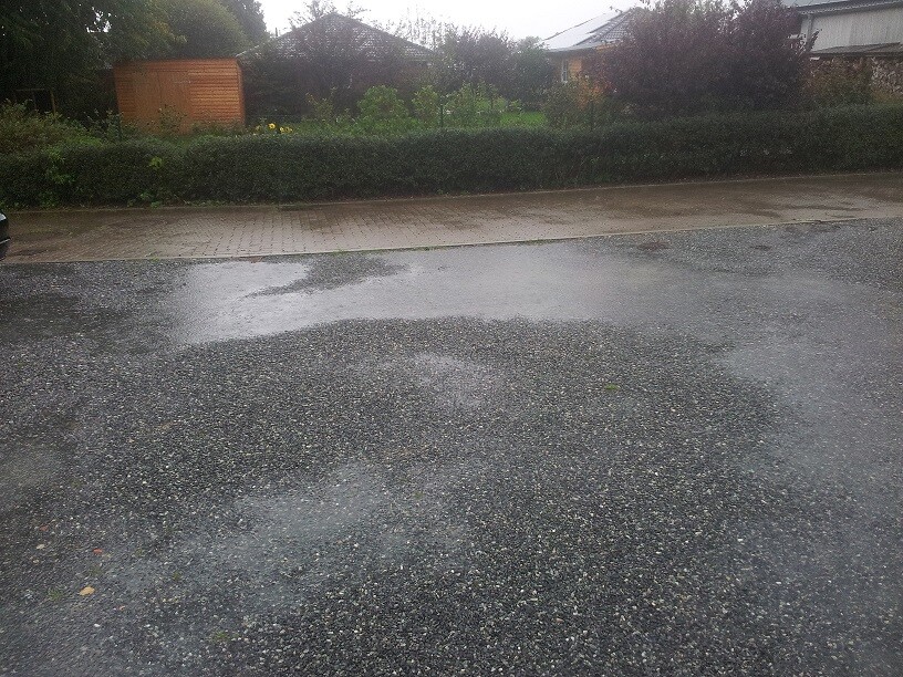 Nasser Vorplatz mit Pfützen vor Wohnhaus, Hecke und Garten im Hintergrund