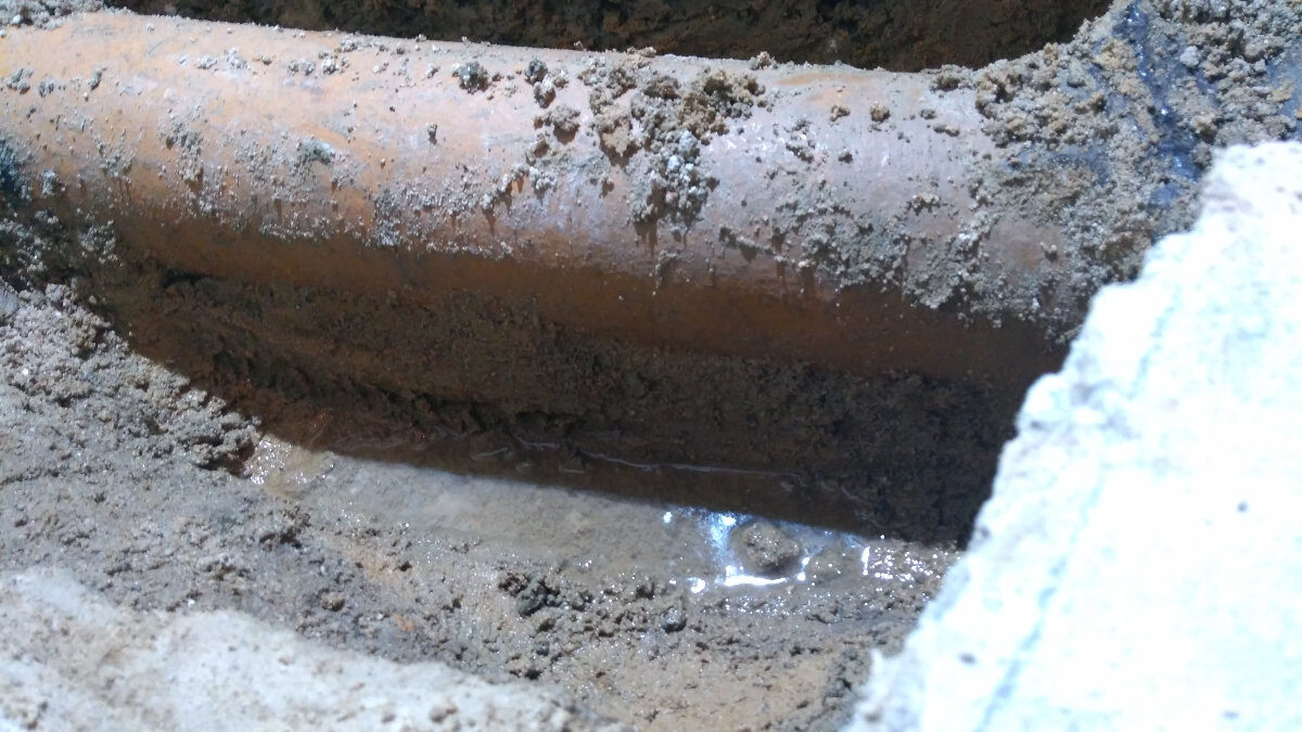Rostiges Rohr im Schlamm, teilweise vergraben, mit nasser Erde und Pfütze.