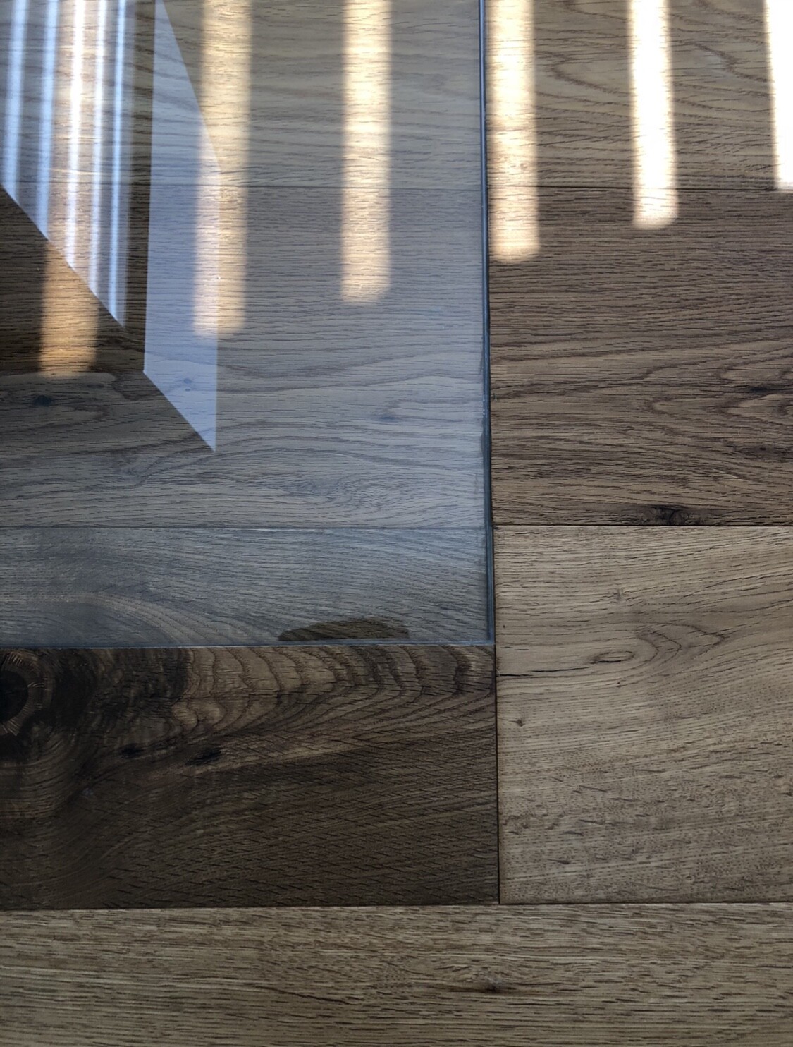 Holzboden mit dunklen und hellen Holzdielen; Glas-Tisch spiegelt vertikale Lichtstreifen.