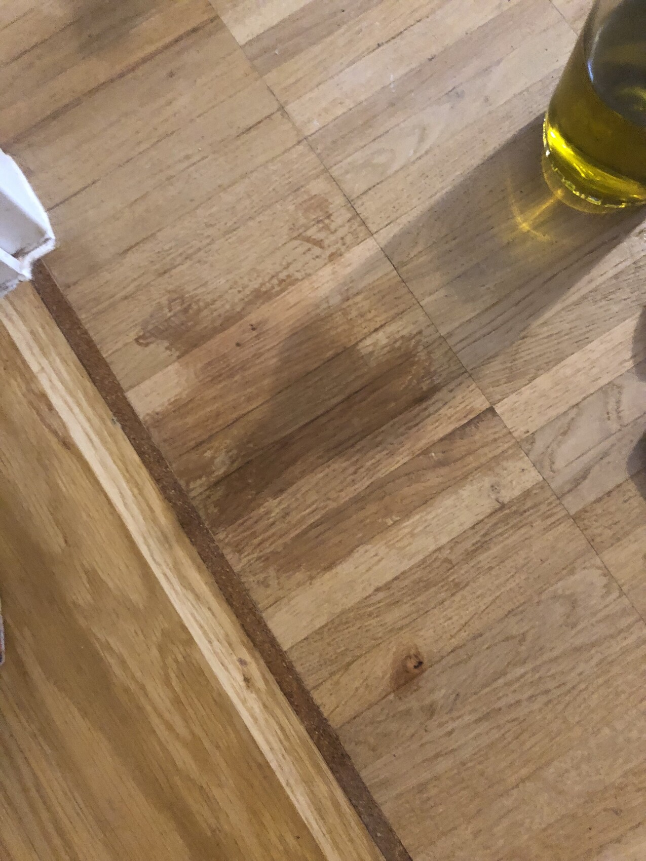 Holzboden mit Bretterverlauf; rechts steht eine Glasflasche mit gelber Flüssigkeit.