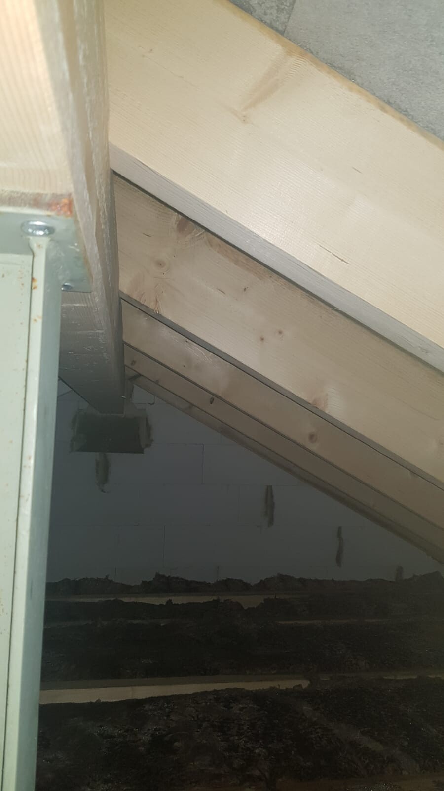 Schräger Dachstuhl aus hellen Holzbalken, links Metallhalterung, Betonwand, dunkler Boden.
