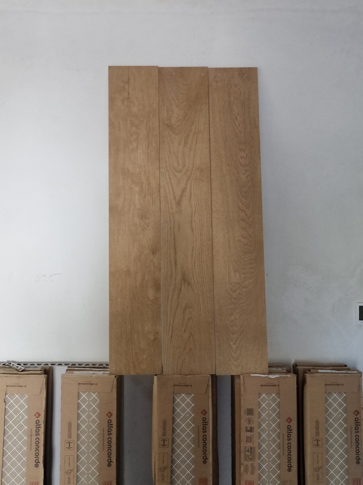 Vier lange Holzbretter mit Holzmaserung stehen auf Kartonverpackungen vor einer hellgrauen Wand.
