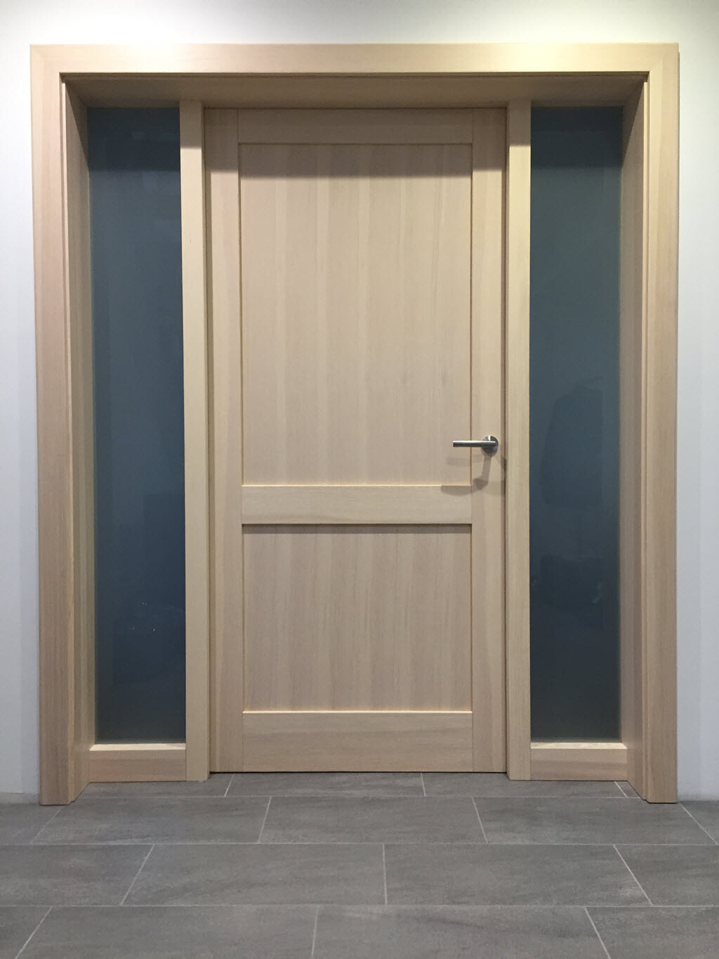 Helle Holztür mit vertikalen Paneele, flankiert von grauen Glasfenstern.