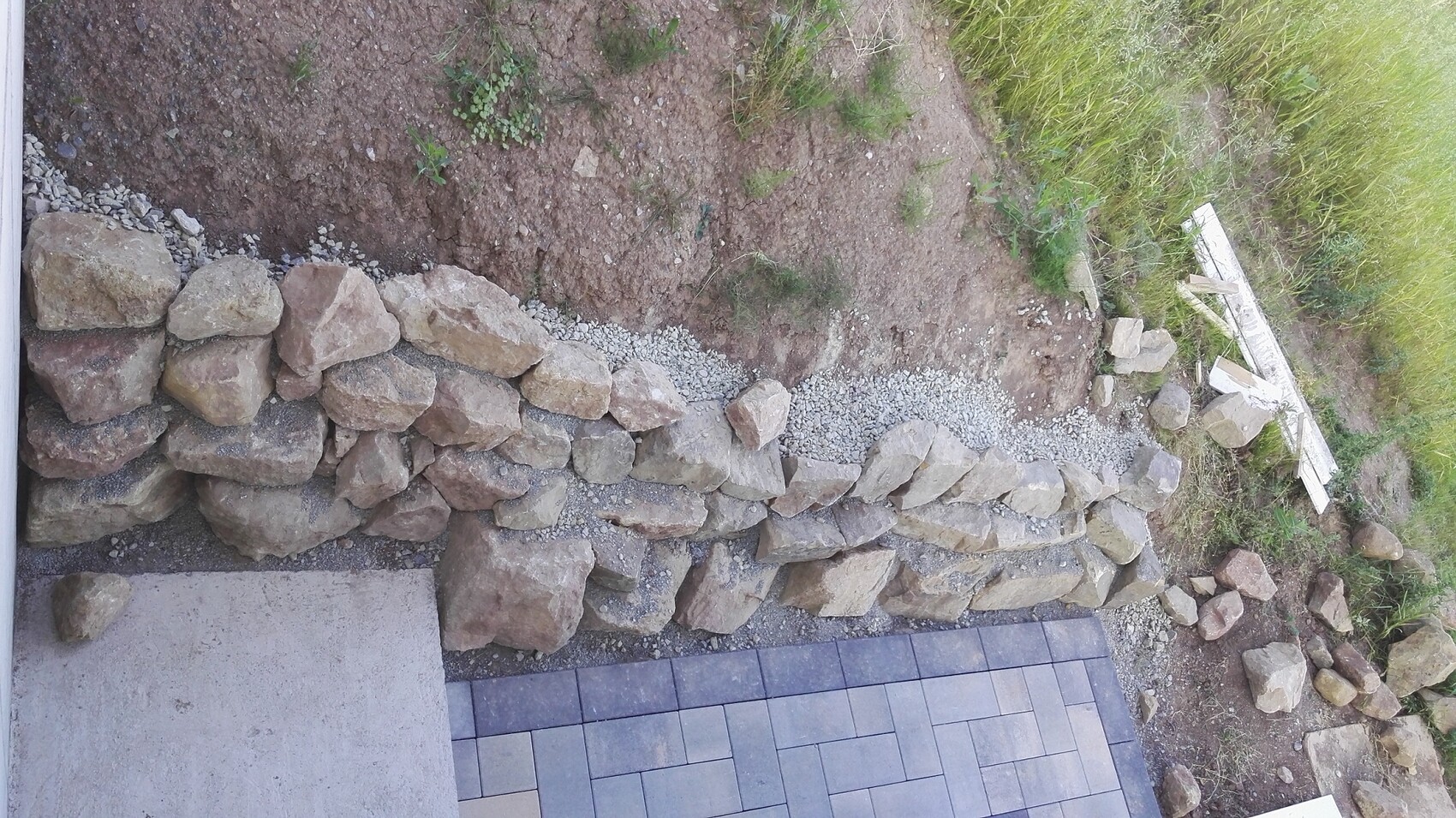 Unregelmäßige Steinmauer aus großen Felsen neben Erdhang; gepflasterte Fläche im Vordergrund.