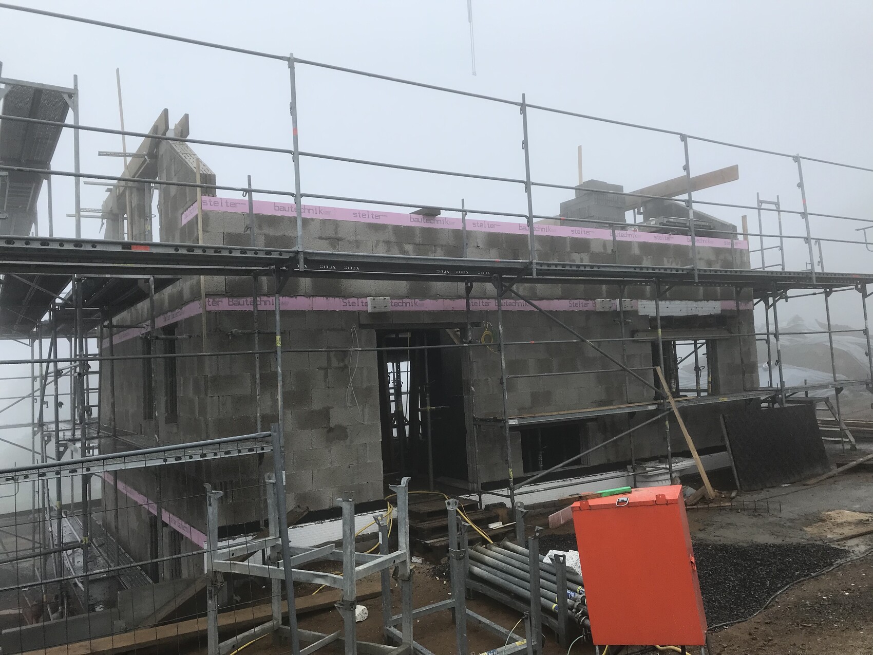 Baustelle mit Gerüst um Rohbau aus Beton, offenem Eingang, pinkes Bauband, orange Kasten.