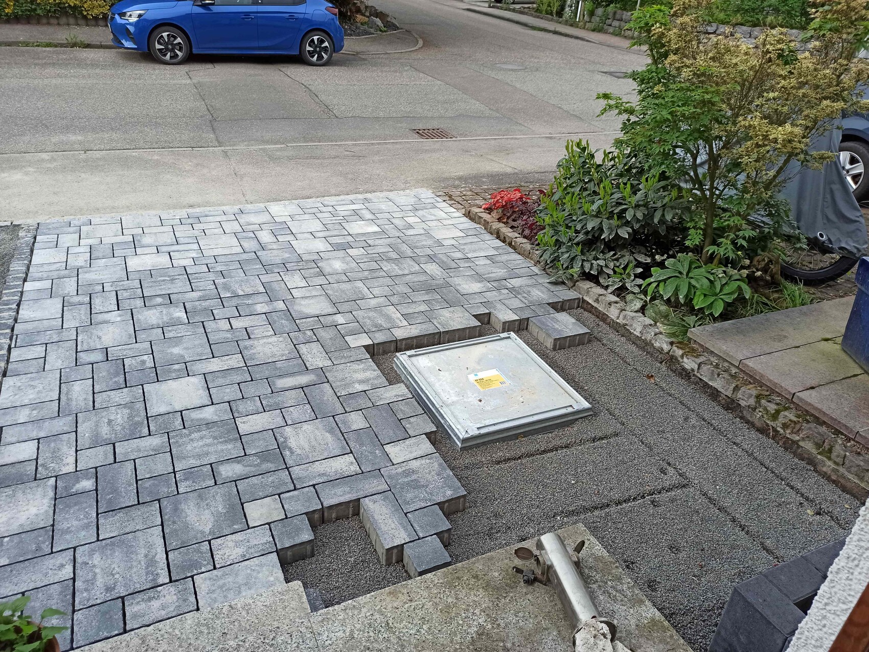 Neu gepflasterter Hofweg mit Metalldeckel, Pflanzenbeet rechts, blaues Auto im Hintergrund.