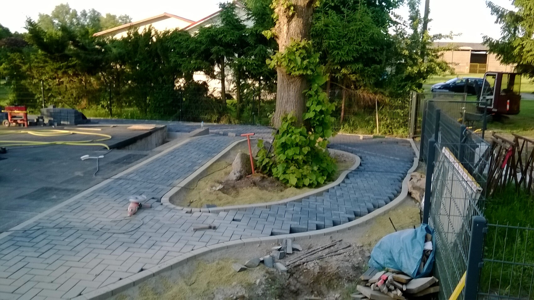Gepflasterter Kurvenweg um Baum mit Kletterpflanze; Bauwerkzeuge und Zaun im Hintergrund.