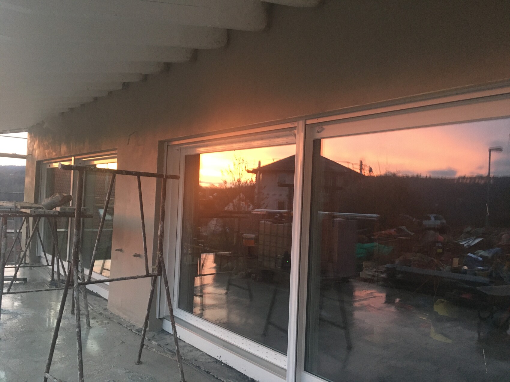 Balkonbaustelle mit Gerüst vor Glas-Schiebetüren; Abendsonne spiegelt sich im Fenster.