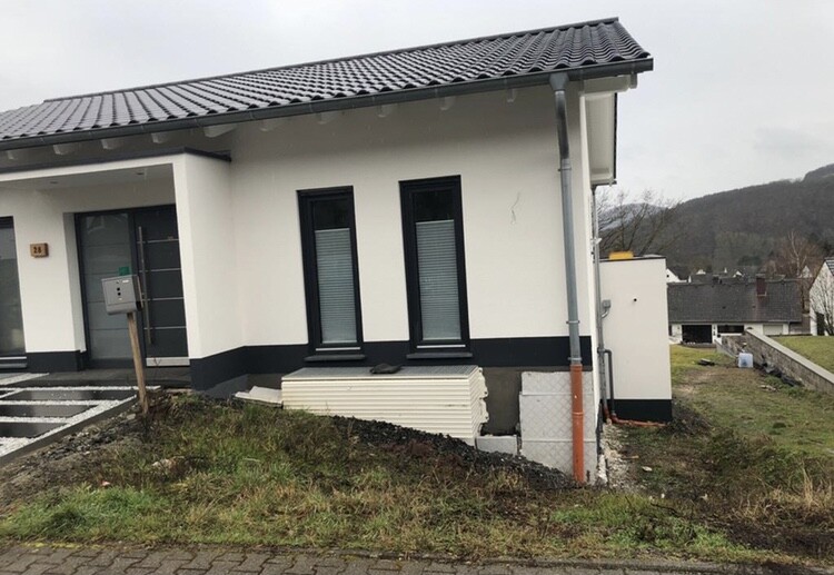 Modernes weißes Einfamilienhaus mit dunklem Sockel, zwei hohe Fenster, Baumaterialien davor.