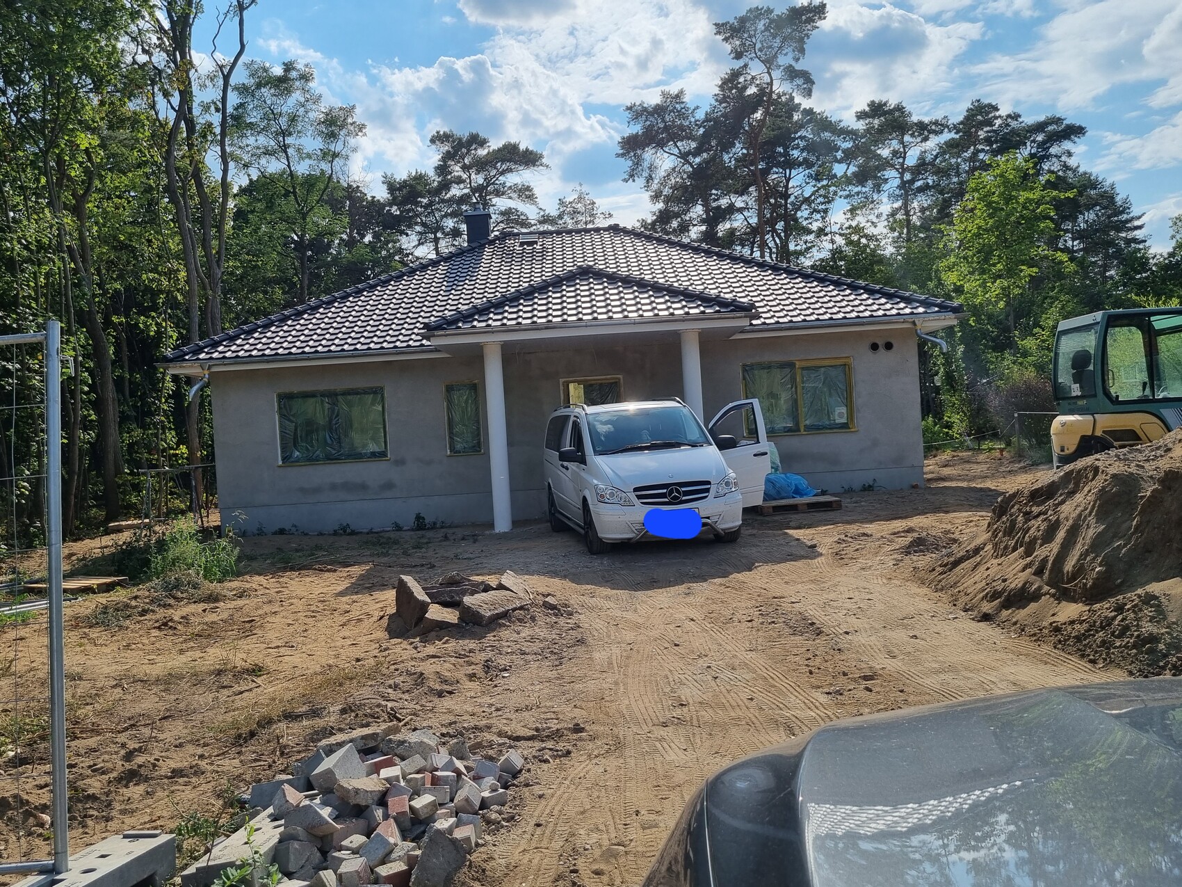 Im Bau befindliches Einfamilienhaus mit weißem Van vor Eingang, Bagger rechts, Wald dahinter.