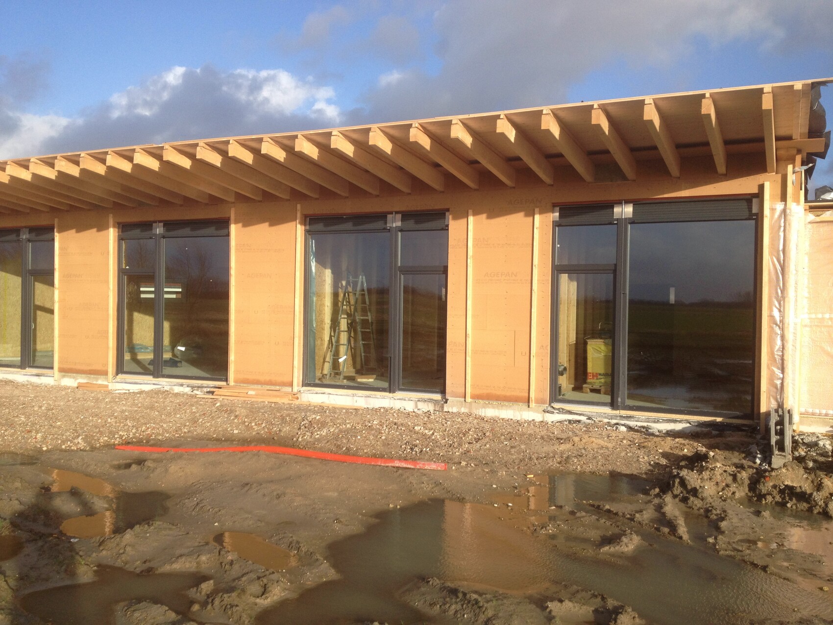 Rohbau eines Gebäudes mit großen Glas-Schiebetüren, Holzrahmen und Bauzone mit Pfützen.