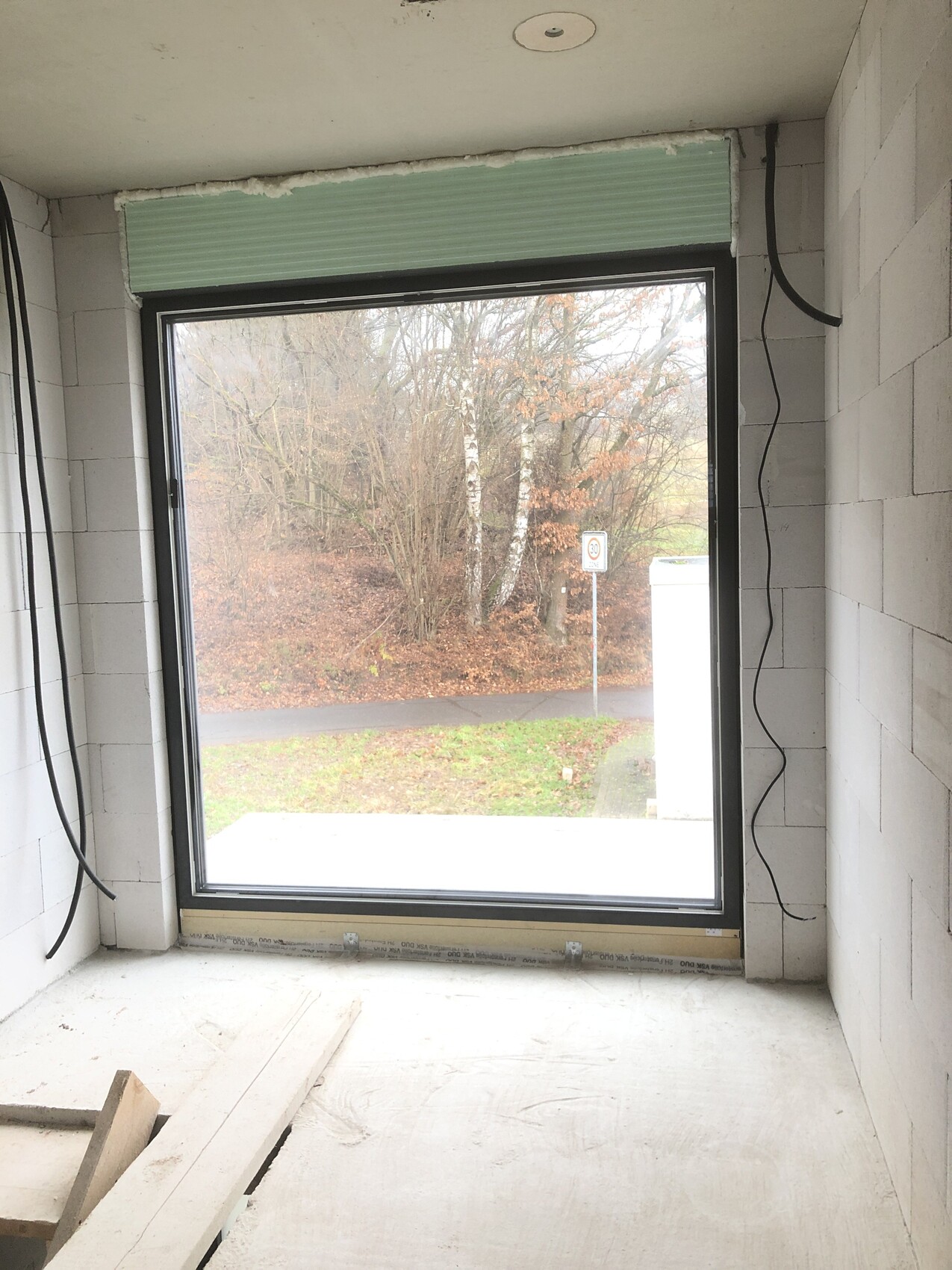 Unfertiger Innenraum mit großem Fenster, Blick nach draußen auf Bäume, Straße und 30er Schild.