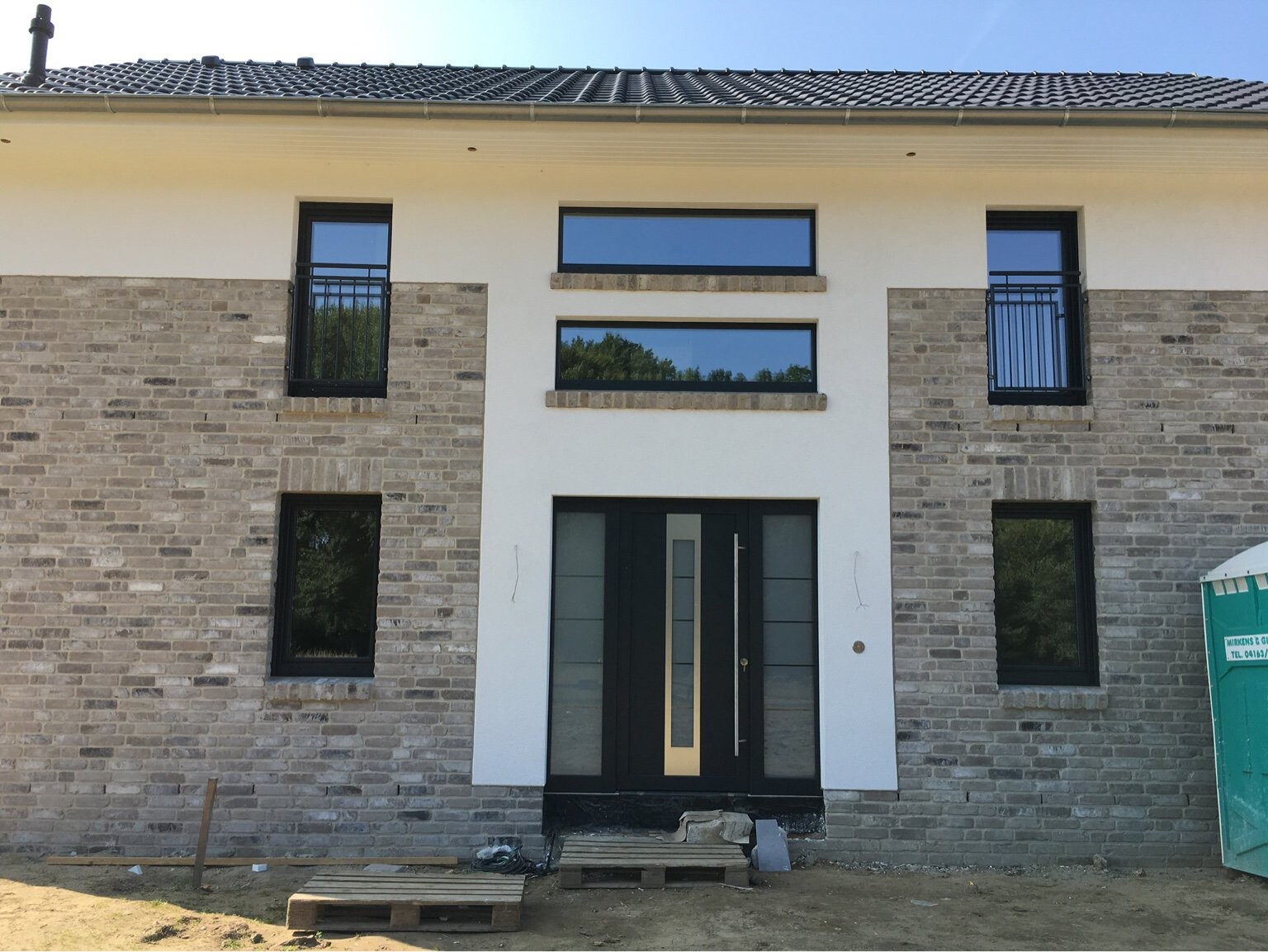 Zweistöckiges Haus im Bau: Steinfassade, weiße Mittelzone mit dunkler Eingangstür, Baupaletten davor.