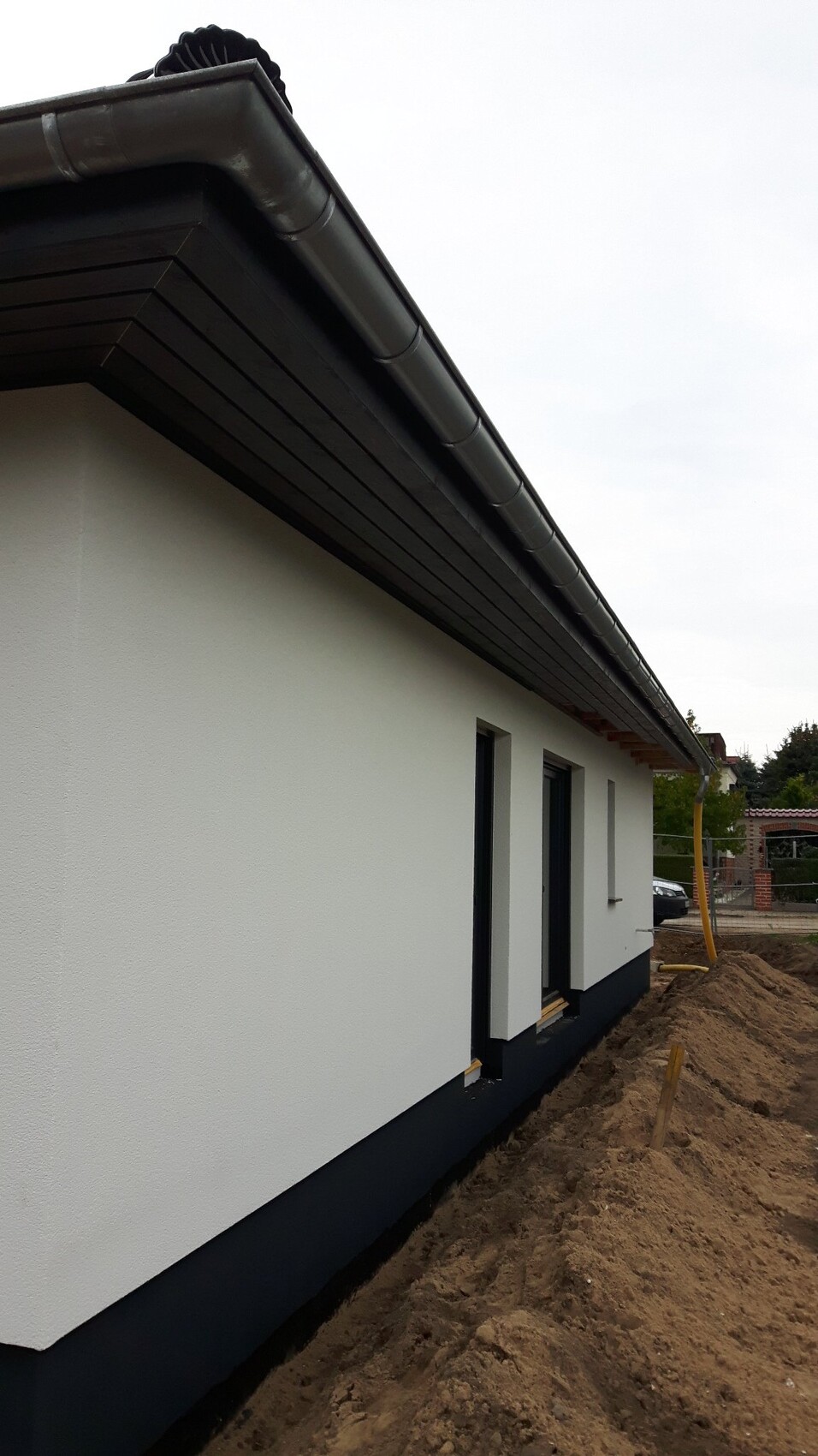 Weiße Hauswand mit schwarzer Dachrinne, Baugrube und Erde am Fundament.