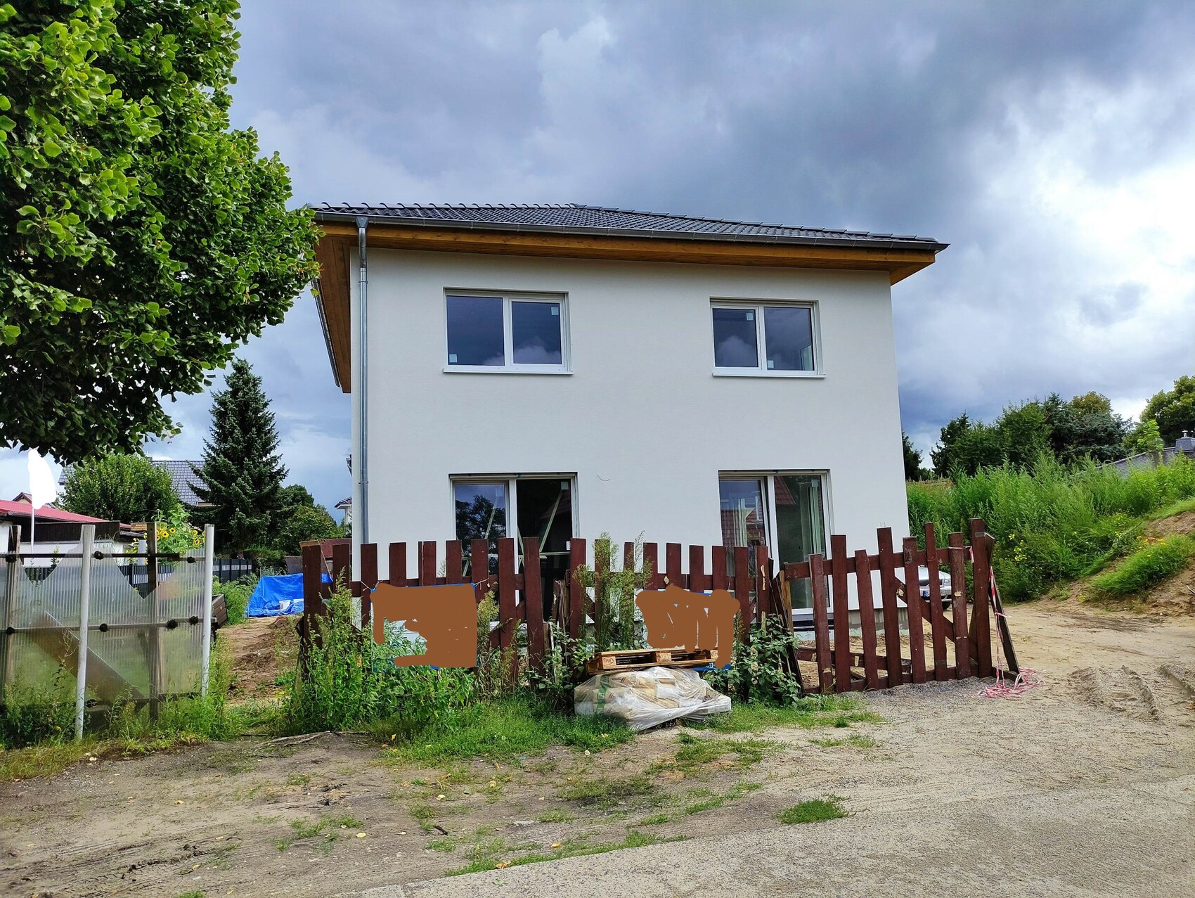Zweistöckiges weißes Haus hinter provisorischem Holzzaun; Bau- und Gartenmaterialien davor.