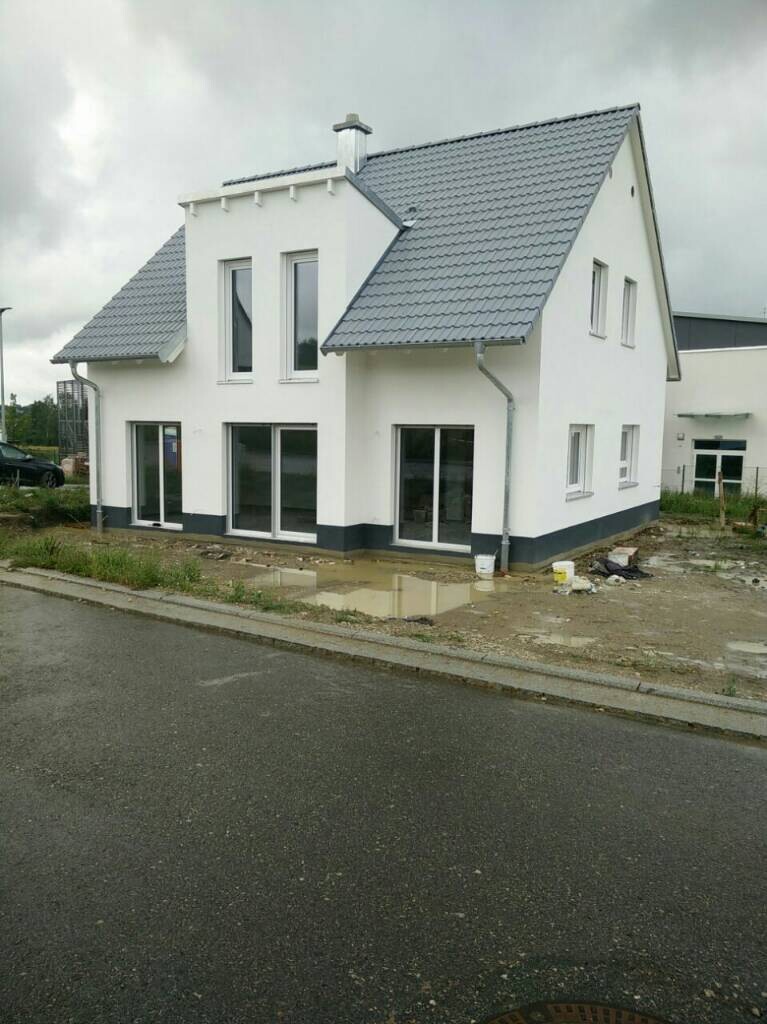 Weißes, zweistöckiges Haus mit grauem Ziegeldach und großen Glasfronten; Baugelände.