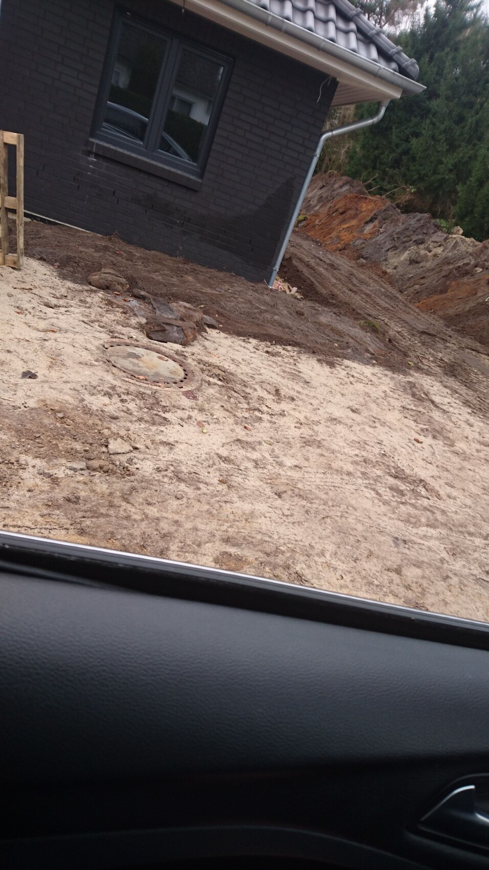 Baustelle neben dunklem Ziegelhaus; Sandboden, Erdhügel, runde Abdeckung, Blick aus Auto.