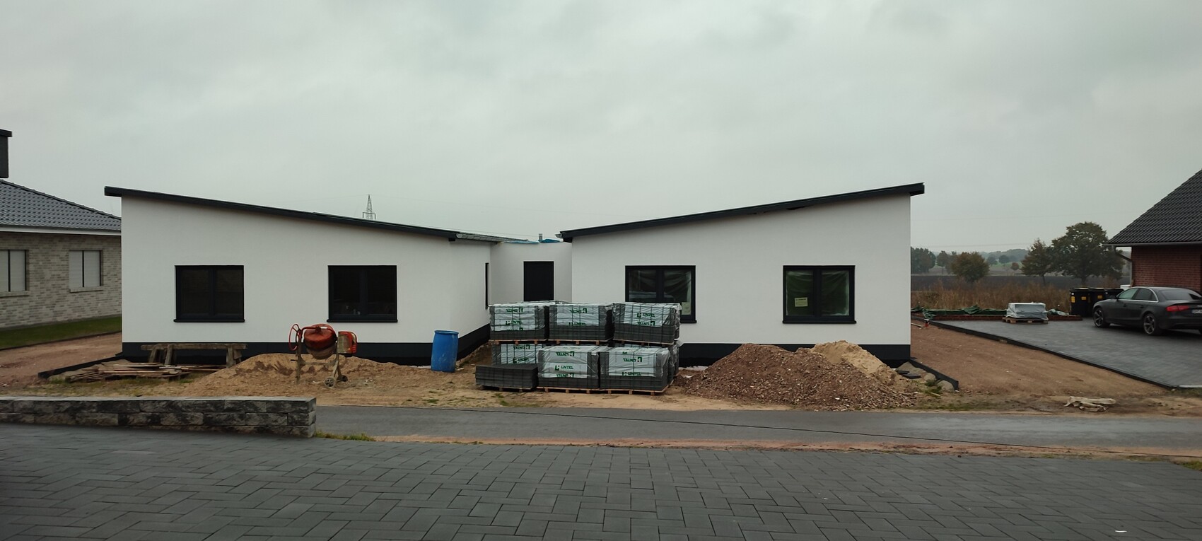 Zwei weiße moderne Bauhäuser auf einer Baustelle mit Betonmischer, Materialpaletten und Sandhügel.