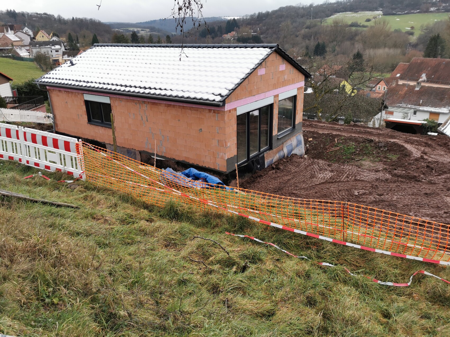 Rohbau eines Hauses auf Hanglage, Schnee auf dem Dach, orange Bauzäune am Baugelände.