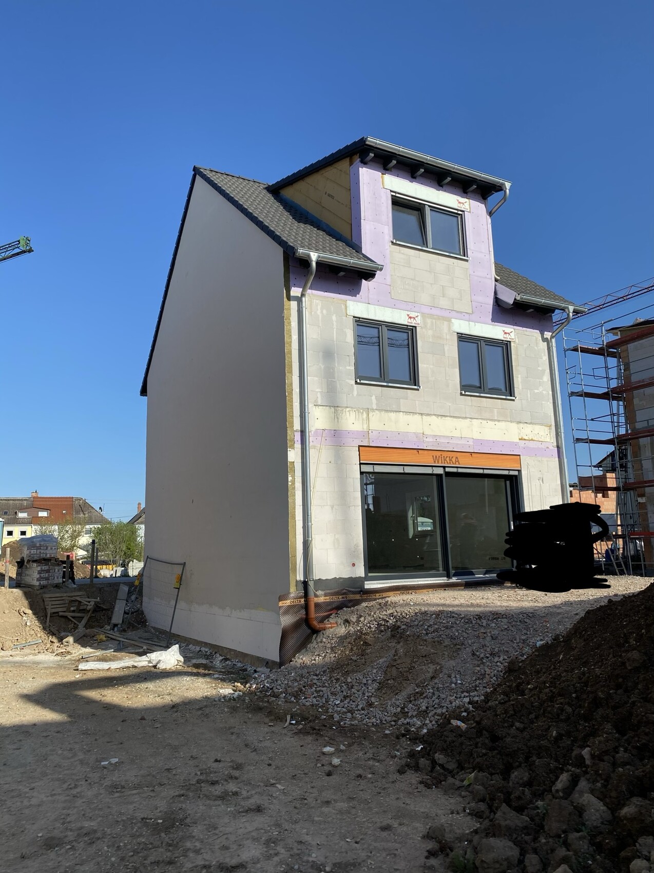 Mehrstöckiges Haus im Bau mit Dämmplatten, Glasfront und Gerüsten rechts.
