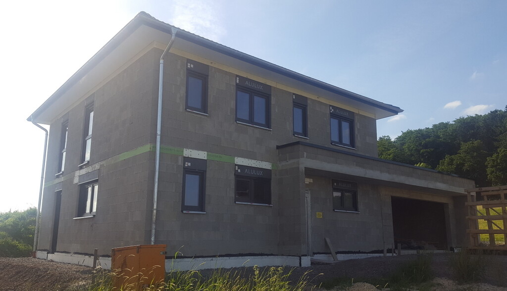 Neubau: zweistöckiges Betonhaus mit offener Garage rechts und dunklen Fensterrahmen.