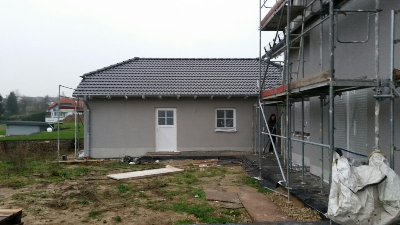 Kleines Haus mit Ziegeldach, rechts Baugerüst, Baustelle mit Materialien, Person vor der Tür.