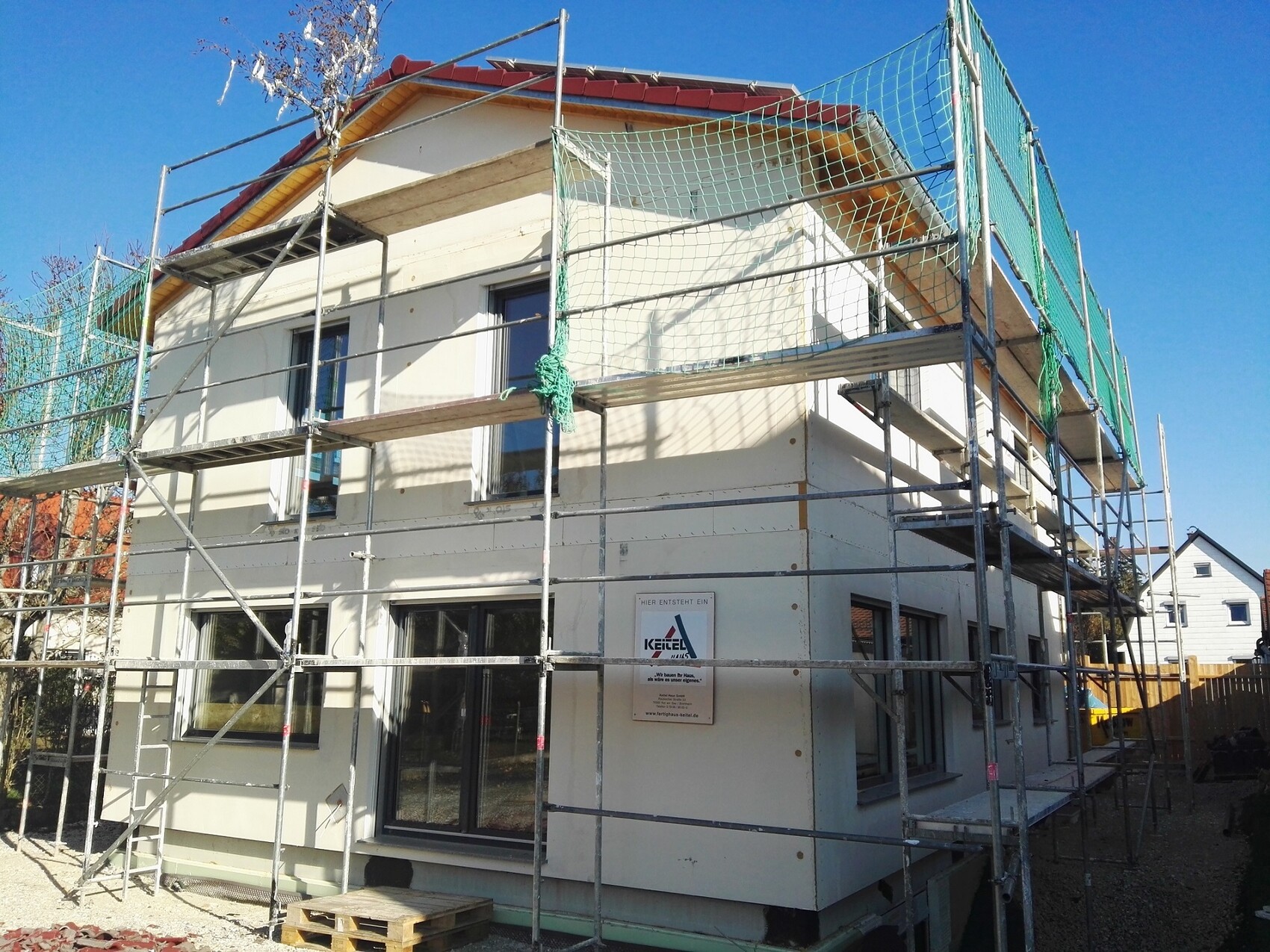 Zweistöckiges Haus im Bau mit Gerüst und grünen Netzen, blauer Himmel.