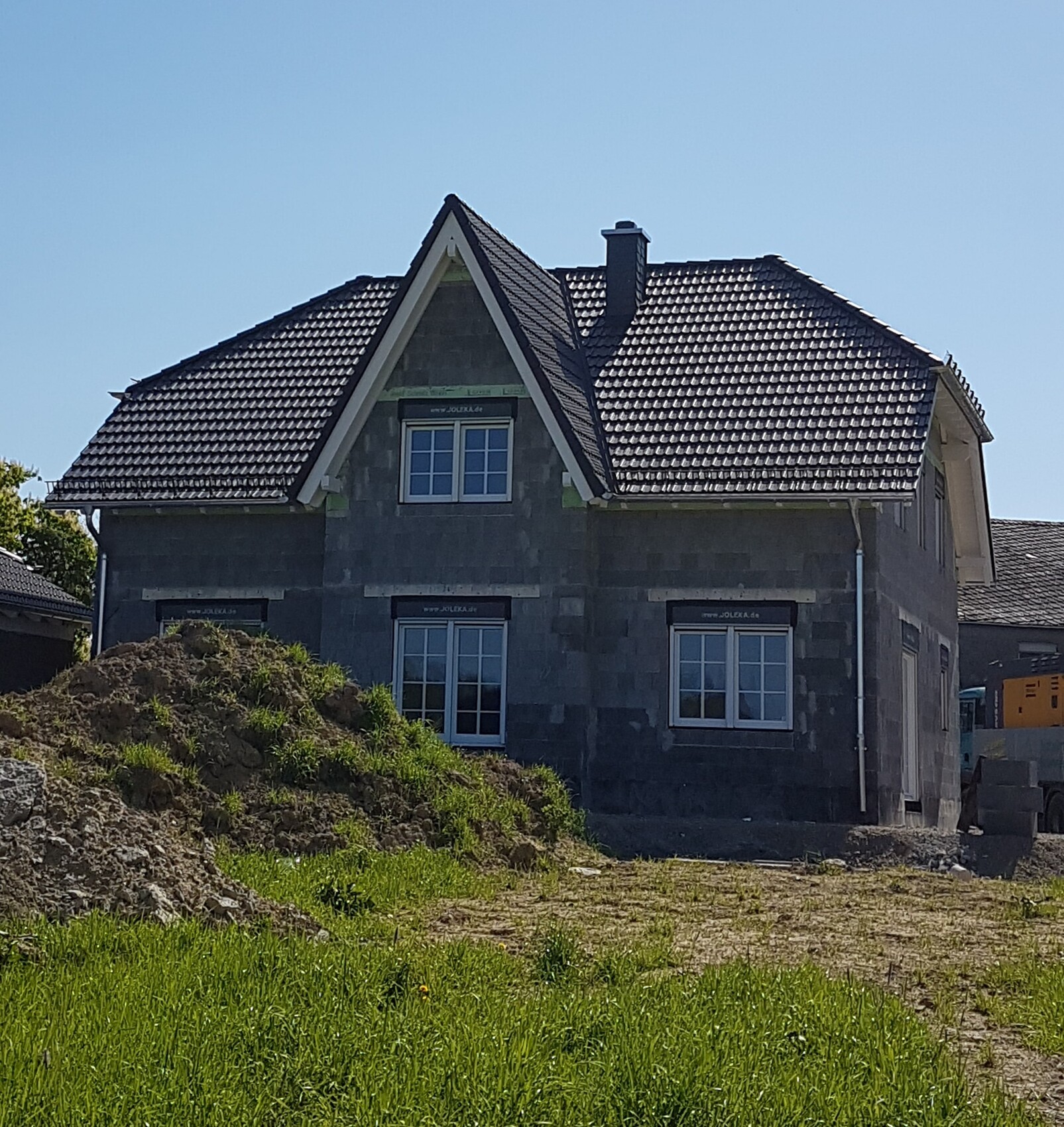 Haus im Rohbau mit dunklem Ziegeldach, Fenstern, Bauhügel vor Grünfläche