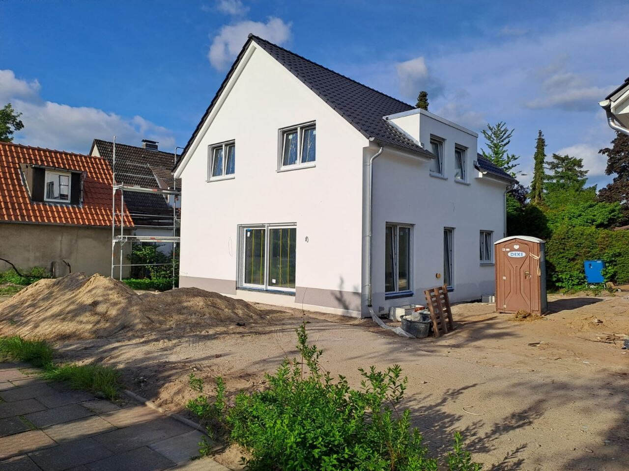Weiße Neubau-Baustelle mit Sandhaufen, Baugeräten und Porta-Potti im Vordergrund